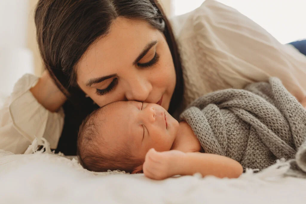 Woman lovingly kisses the forehead of a sleeping newborn wrapped in a gray blanket on a bed, capturing a tender moment of newborn care during daytime care postpartum.