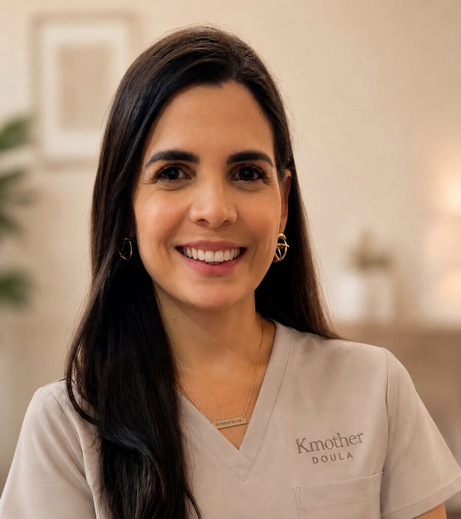 Smiling woman with long dark hair wearing scrubs that say "Kmother Doula," standing indoors, ready to tell you about Kmother Doula and how we support families.